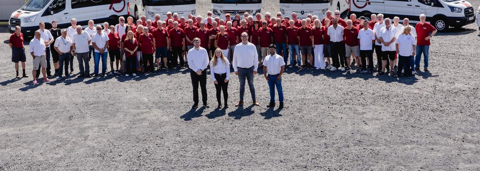 Teamfoto 2026: Belegschaft von Dornburg Reisen in roten und weißen Poloshirts vor Reisebussen und Kleinbussen auf dem Betriebshof