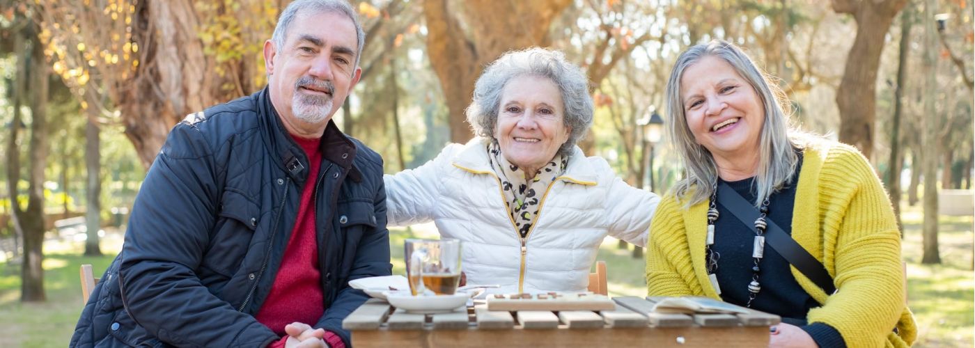 Drei ältere Erwachsene sitzen lachend an einem Holztisch in einem herbstlichen Park